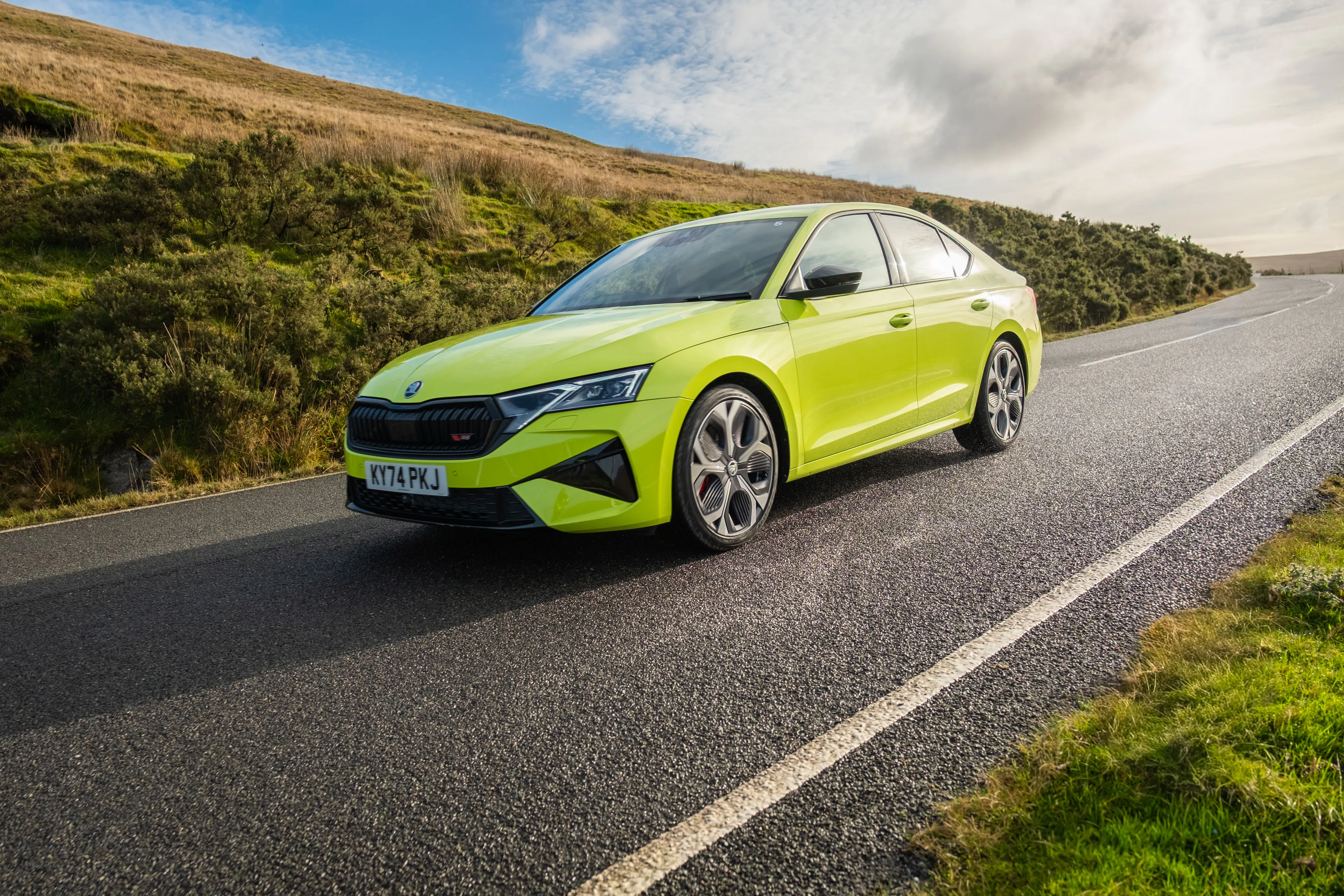 Bright green Skoda car driving on a winding rural road, surrounded by hills and grass under a partly cloudy sky, conveying a sense of adventure.